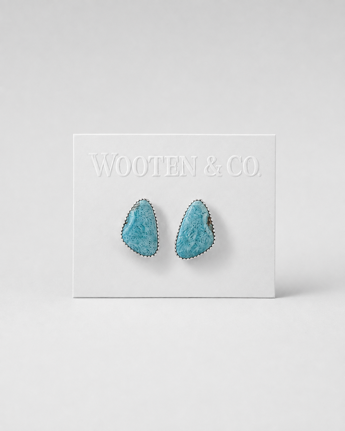 A pair of turquoise-colored stone stud earrings with silver-toned borders, displayed on a white Wooten & Co. brand card against a light gray background.