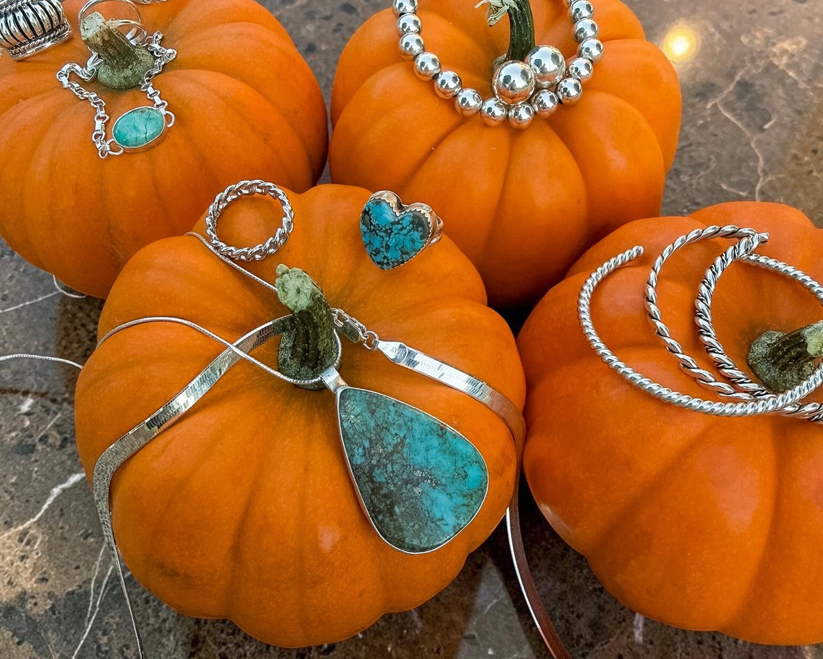 Four orange pumpkins arranged on a brown marble countertop, each decorated with silver-toned jewelry pieces including rings, bracelets, and a teardrop pendant with turquoise-colored stones.