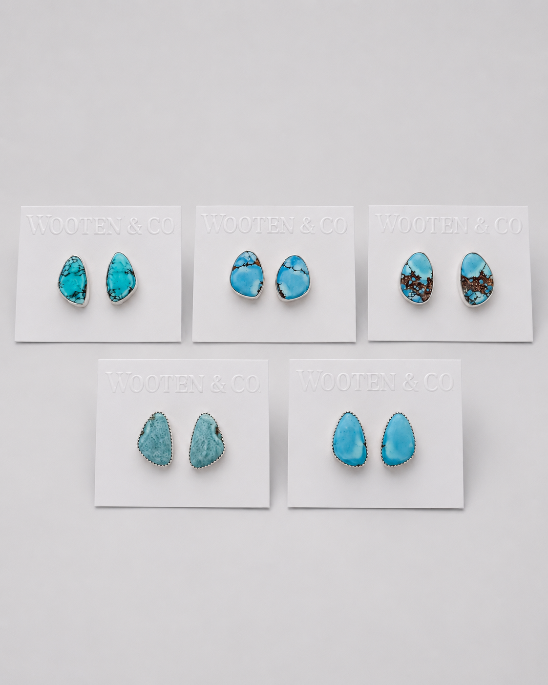 Five pairs of turquoise stud earrings displayed on white backing cards against a light gray background. The earrings feature various turquoise shapes, including oval, teardrop, and irregular natural stone forms, with some showing dark brown matrix patterns.
