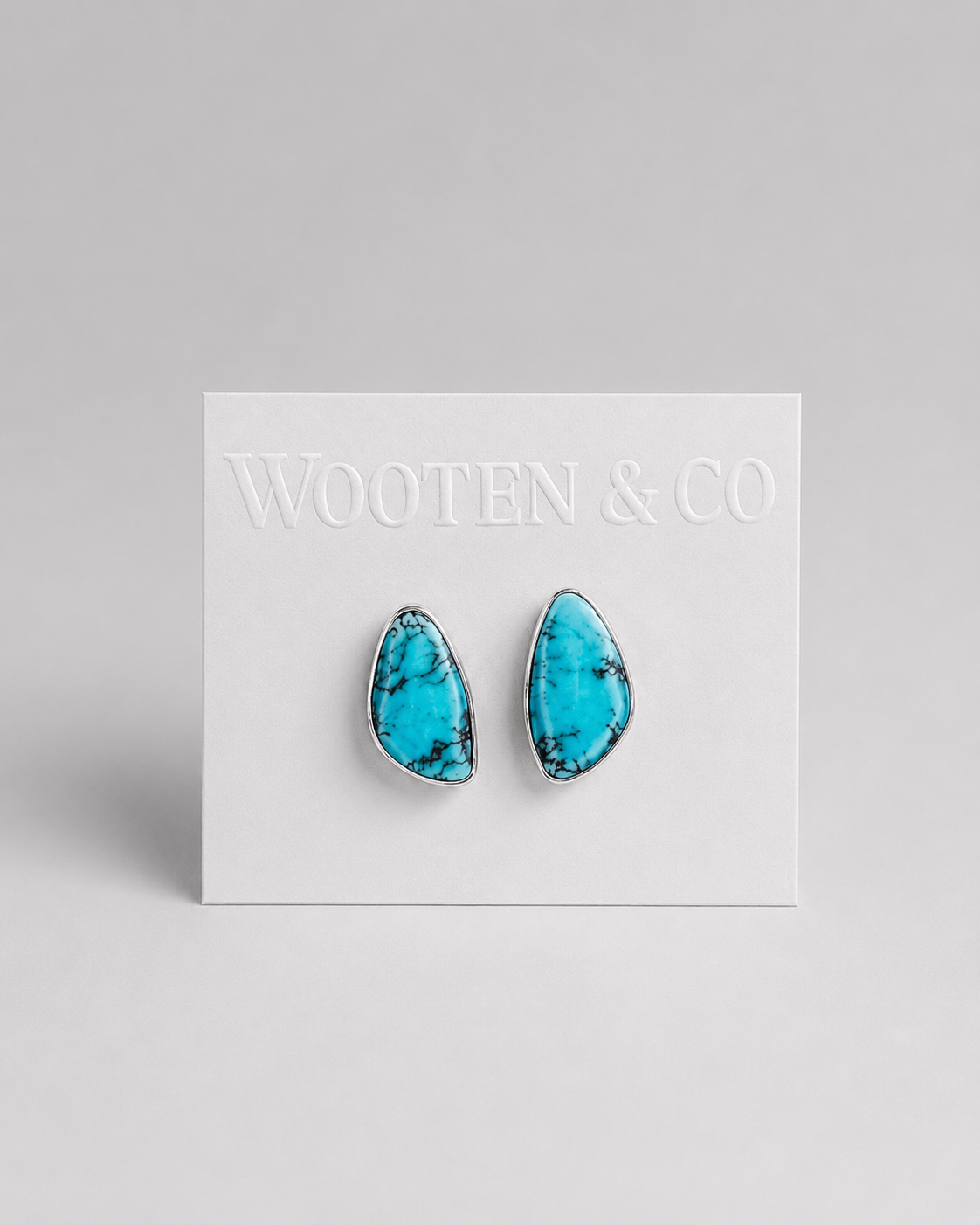 A pair of Wooten & Co stud earrings with turquoise blue marbled stones in silver bezel settings, mounted on a white display card against a light gray background.