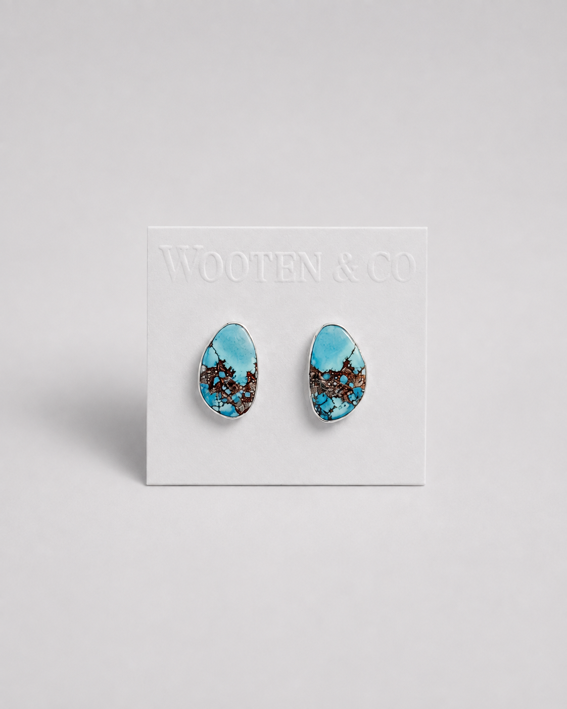 A pair of Wooten & Co. stud earrings with turquoise blue and brown marbled patterns, mounted on a white display card against a light gray background.