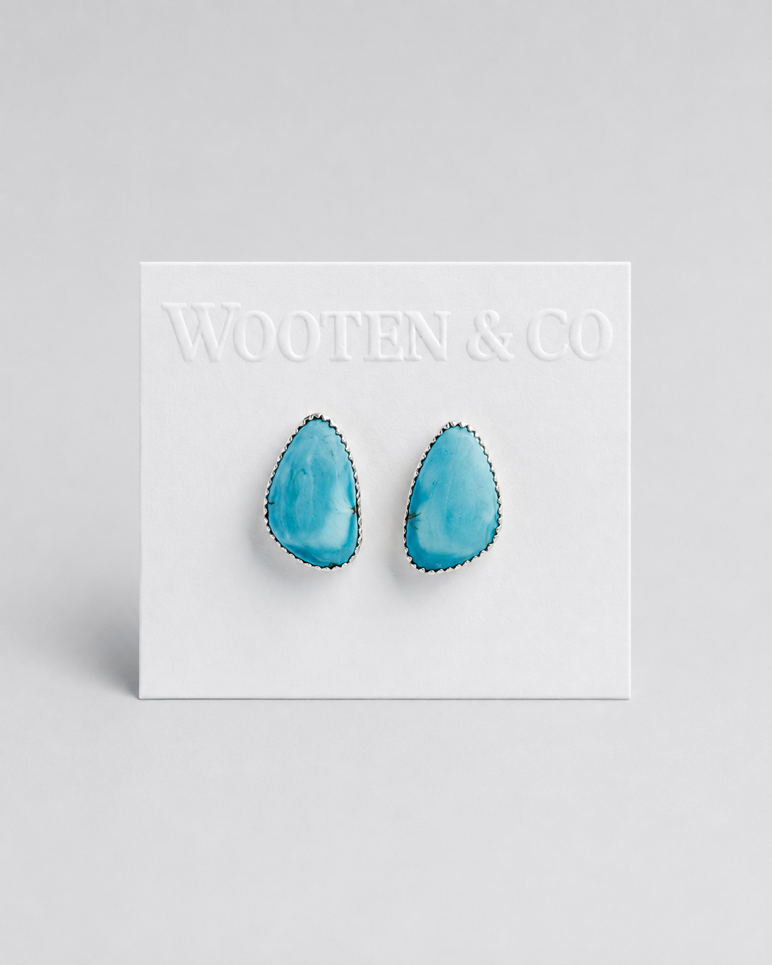 A pair of Wooten & Co stud earrings with turquoise-blue teardrop-shaped stones set in silver, mounted on a white display card against a light gray background.