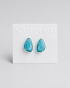 A pair of Wooten & Co stud earrings with turquoise-blue teardrop-shaped stones set in silver, mounted on a white display card against a light gray background.