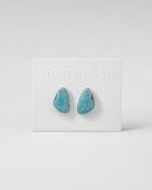 A pair of turquoise-colored stone stud earrings with silver-toned borders, displayed on a white Wooten & Co. brand card against a light gray background.